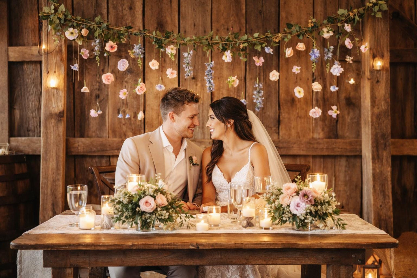 Couple sitting at a sweetheart table decorated with flowers and candles in a rustic wedding setting.
