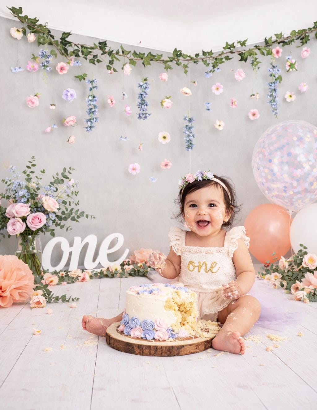 Baby celebrating a first birthday with a cake, balloons, and handmade floral backdrop decorations.