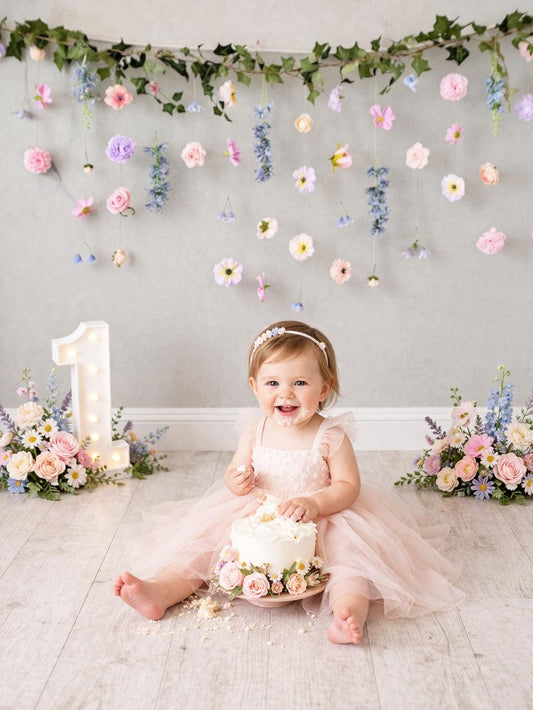 Child in a pink dress sitting on the floor with a small cake, surrounded by floral decorations and a number one candle at cake smash photoshoot.