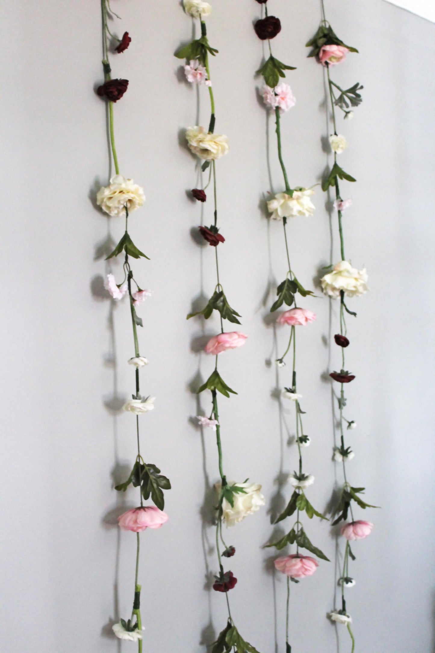 Three long hanging garlands of artificial flowers with pink, cream, and burgundy blossoms and green stems, displayed against a plain light gray wall.